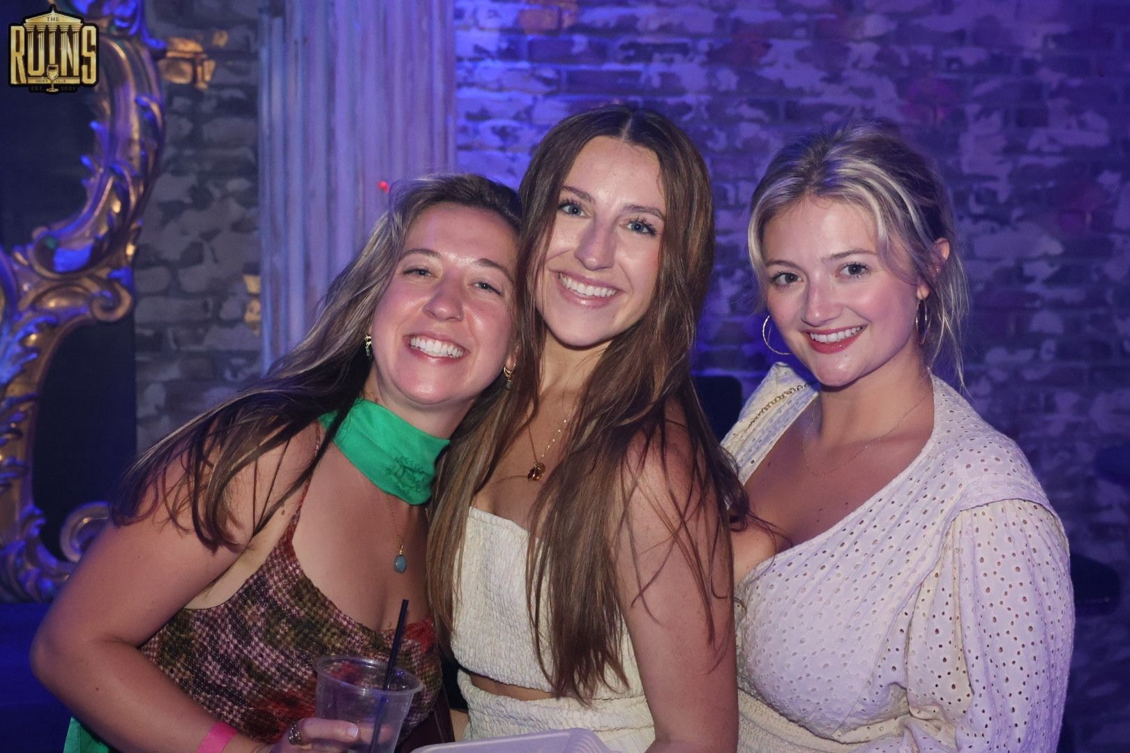 Three friends smiling in a purple-lit club, posing close together at a party.