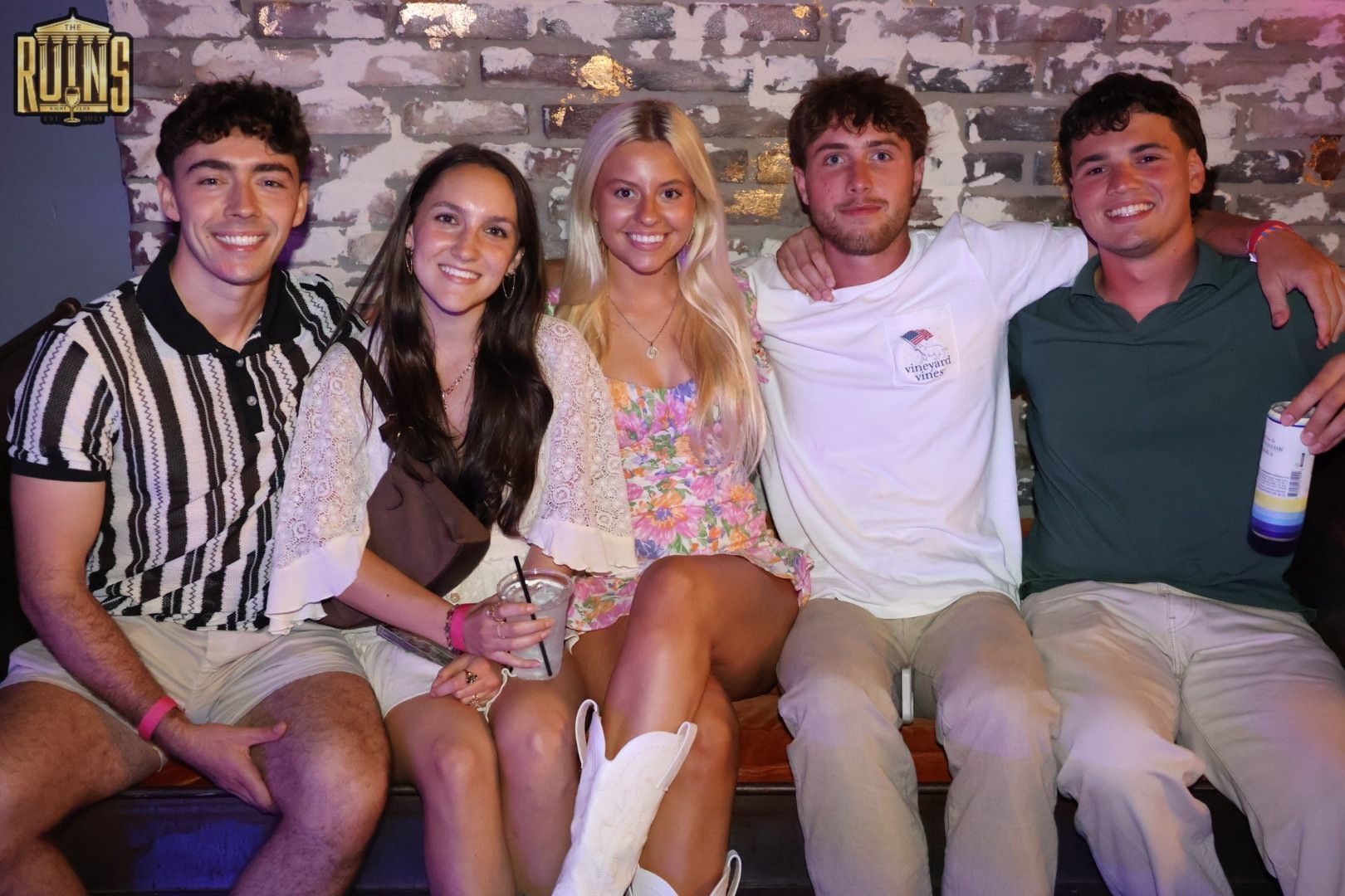 Five friends sit on a couch, smiling at the camera in a club-like setting.