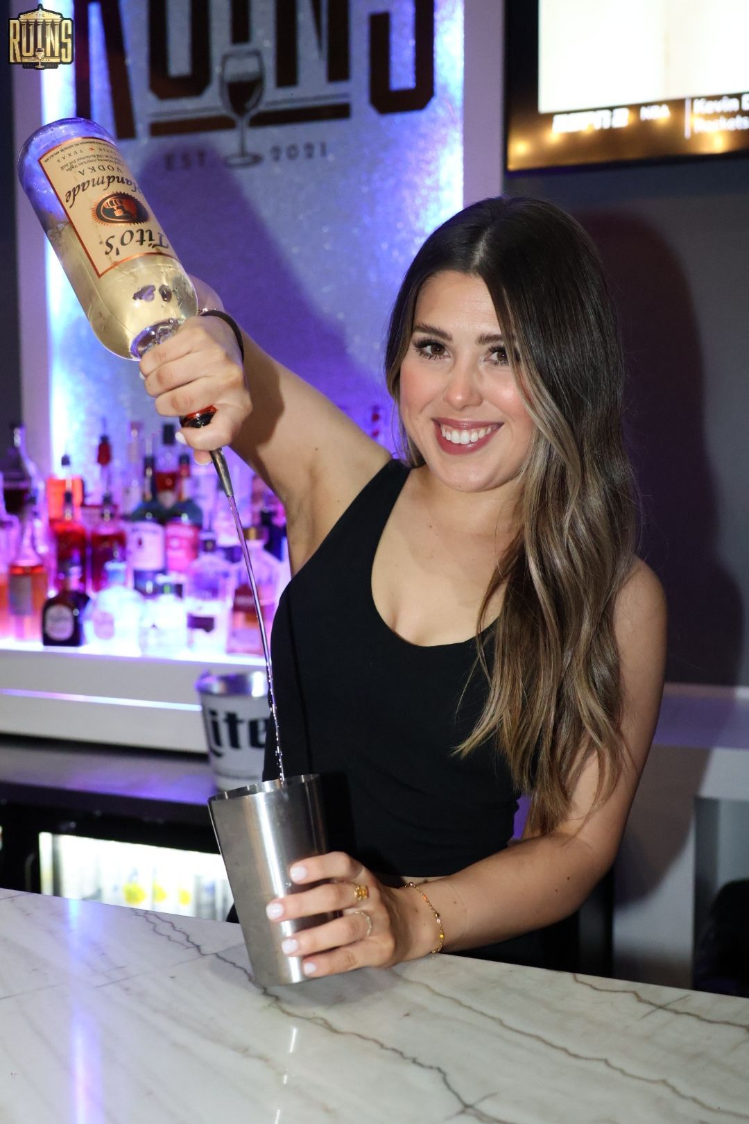 Bartender pouring liquor into a metal shaker at a bar, smiling, with purple lighting in the background