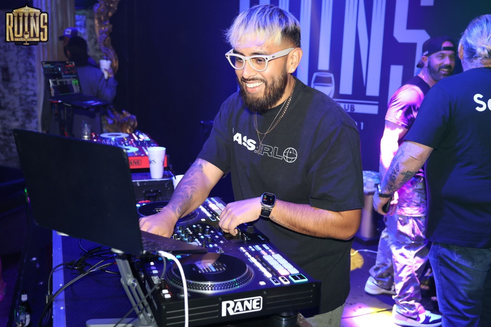 DJ smiling at a turntable in a club, with a laptop and colorful stage lights