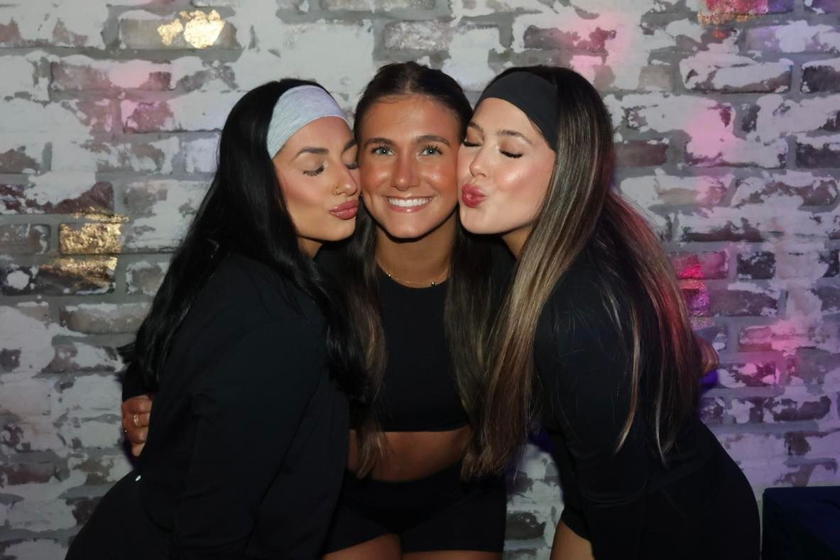 Three friends smiling and posing closely together in black outfits against a brick wall.