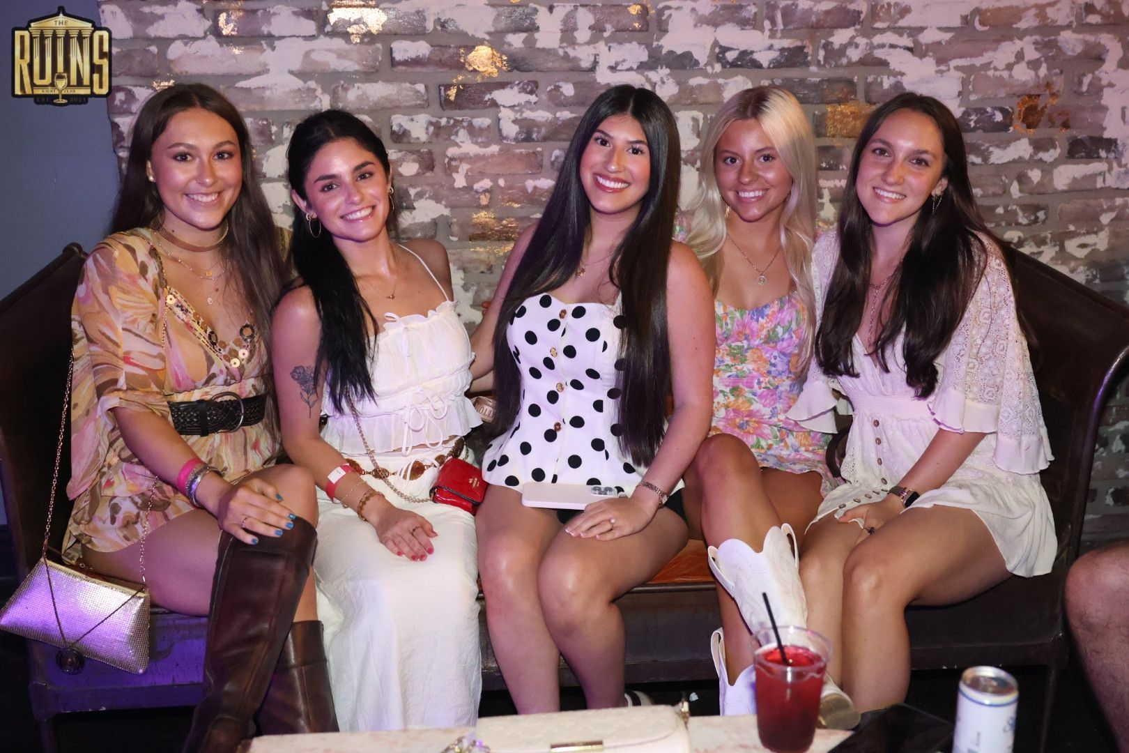 Five friends sit on a couch, smiling at the camera in a dim lounge with drinks on the table.