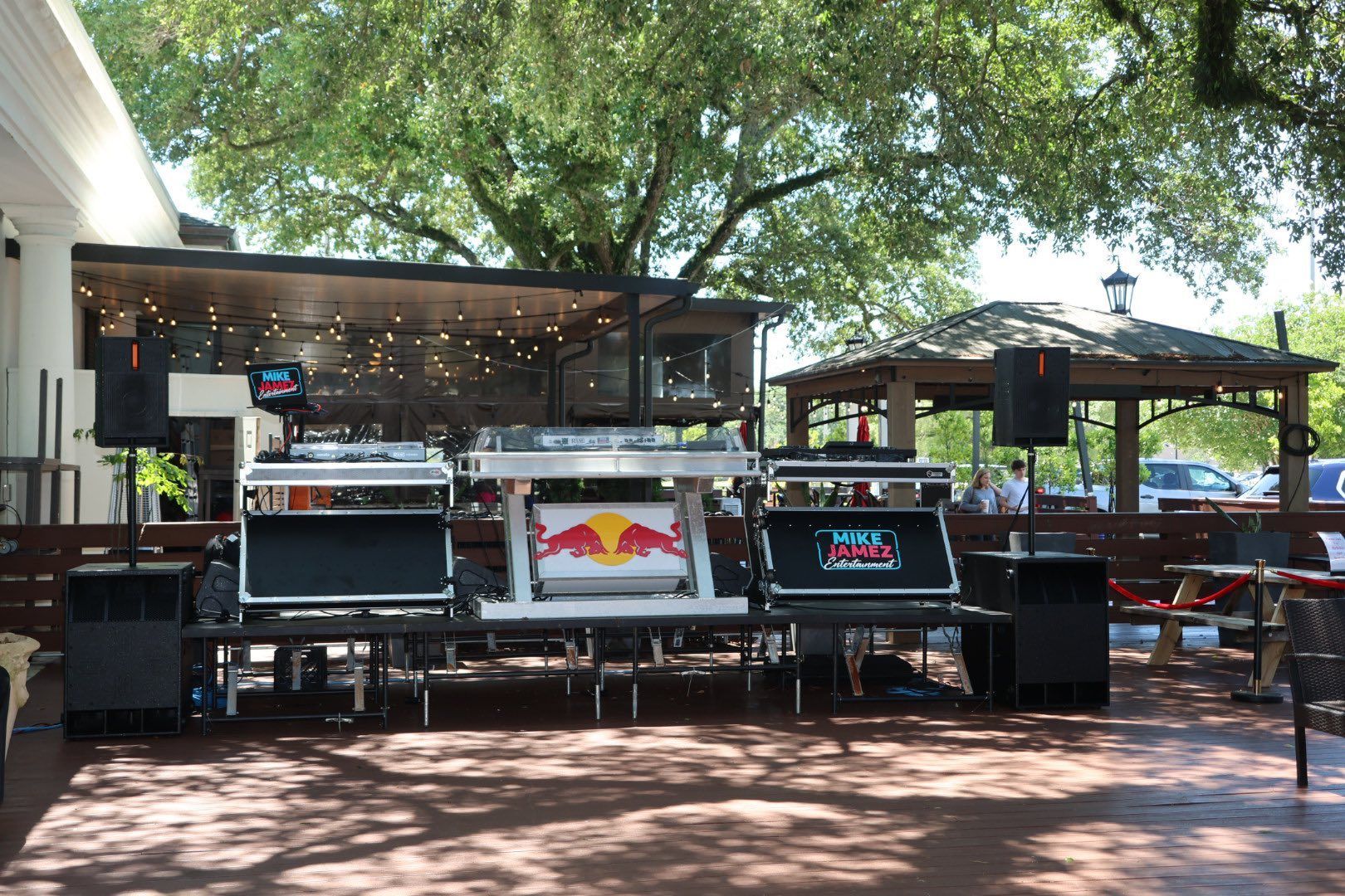 Outdoor live music stage with speakers, red bull sign, and string lights under large trees