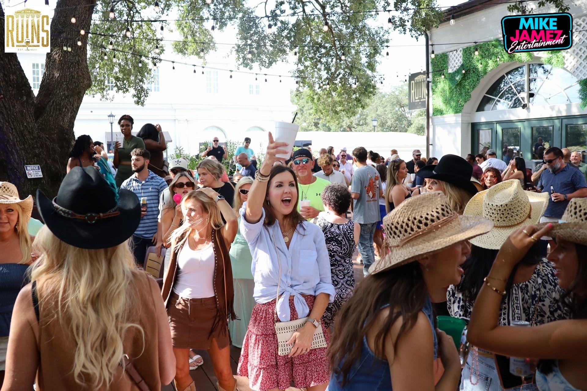Crowd dancing outdoors at a sunny festival, with people in cowboy hats near a tent and trees
