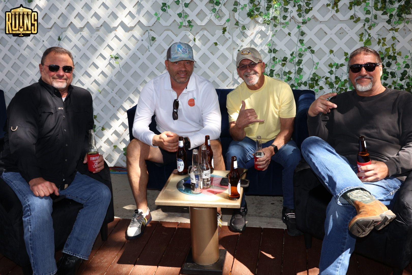 Four men sit on a patio bench by a small table, smiling and holding drinks in bright sunlight.