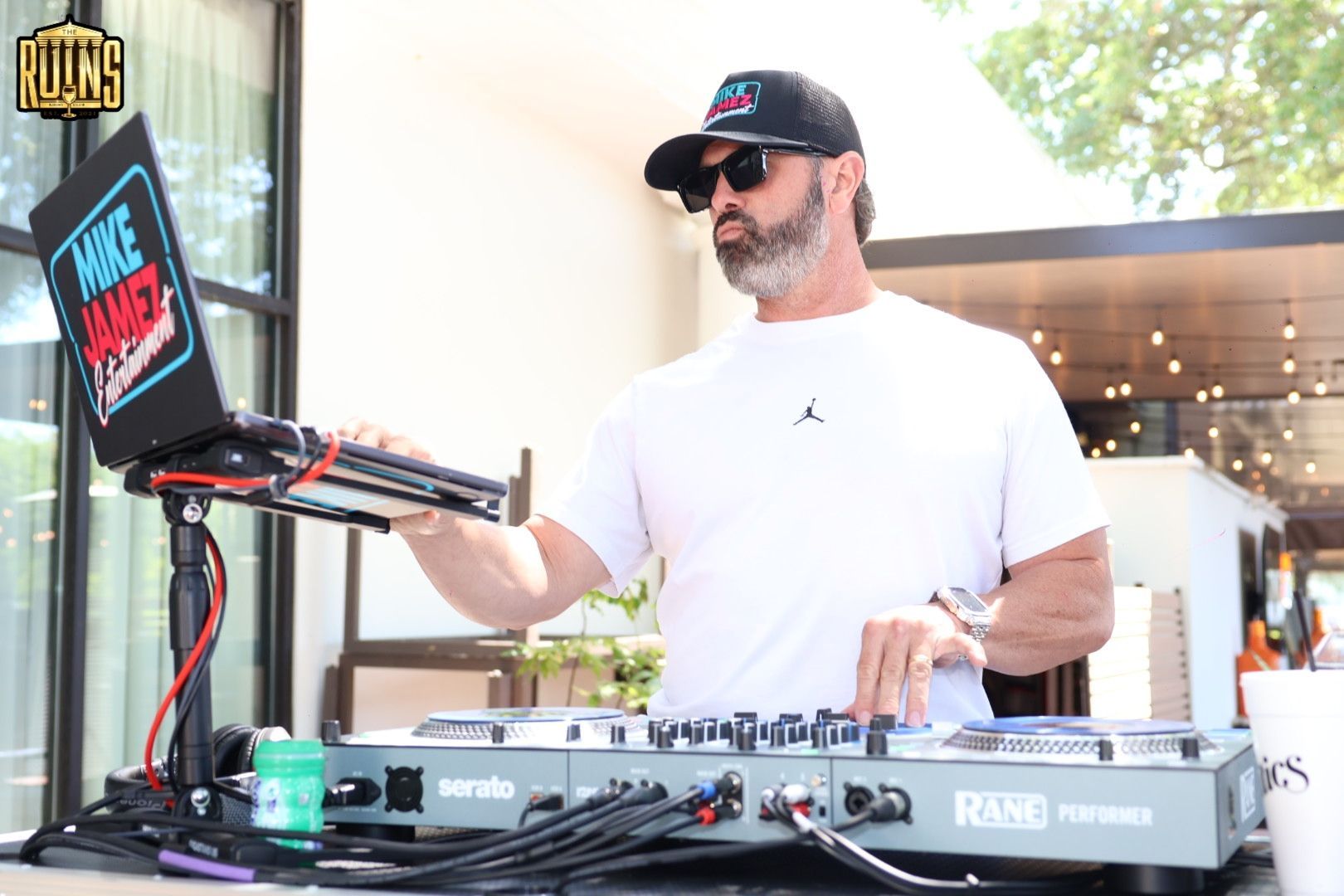 DJ in white shirt and cap mixing music outdoors beside a laptop and turntable setup