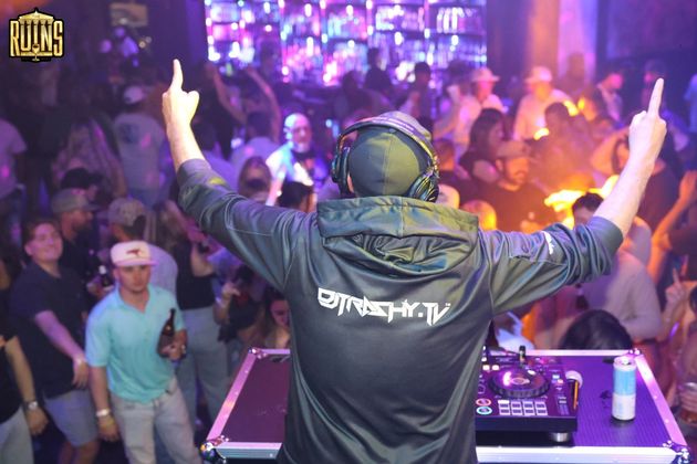 DJ with raised arms performing at a crowded, purple-lit nightclub stage