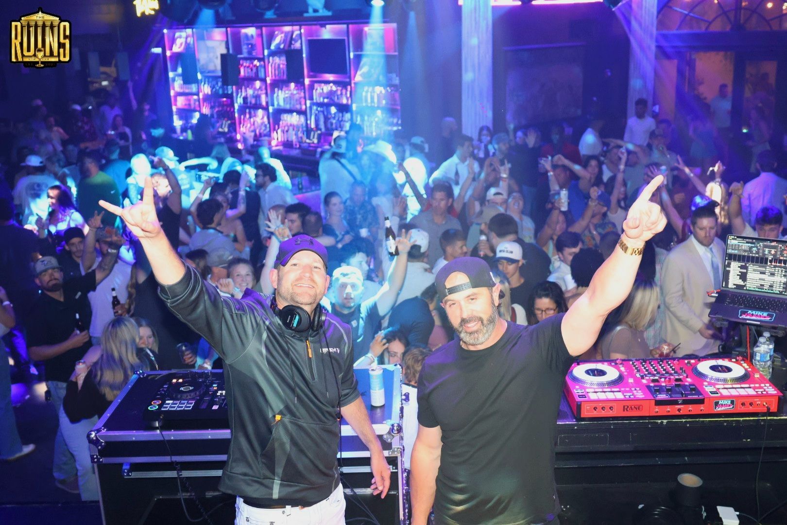 Two DJs smiling and raising their arms at a crowded nightclub booth with glowing lights and turntables