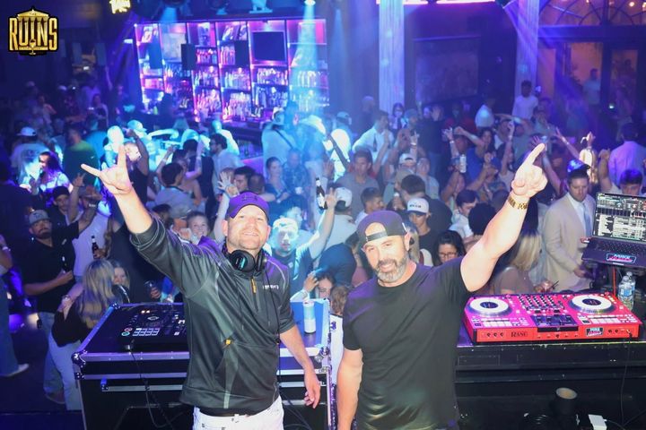 Two DJs posing with raised arms in a crowded nightclub, with blue lighting and DJ equipment in front.