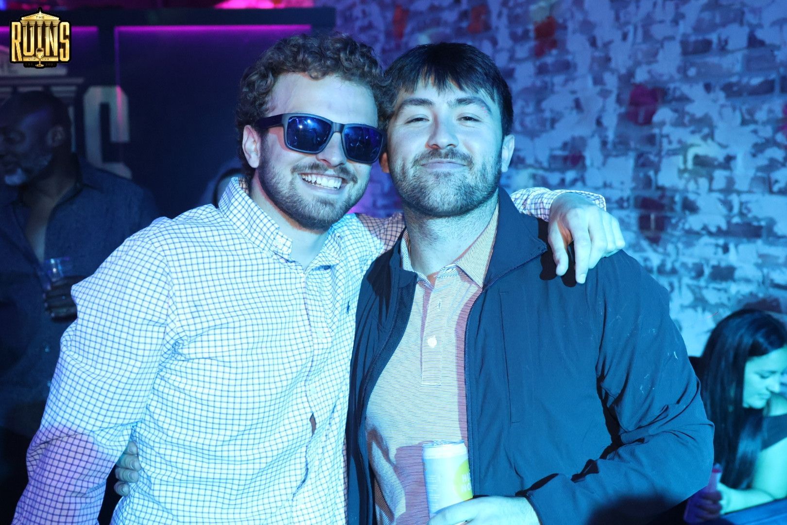 Two friends posing together at a blue-lit party, smiling with arms around each other.