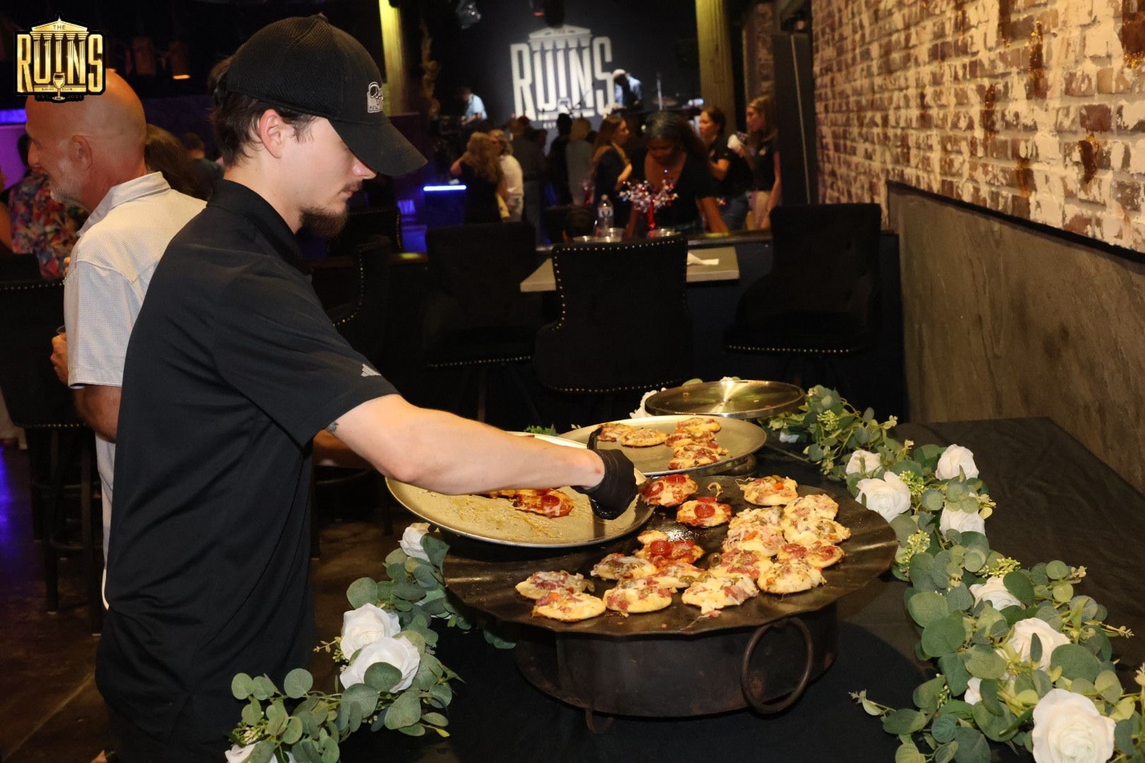 Man serving food at a buffet table in a dimly lit restaurant with flowers and pizza trays