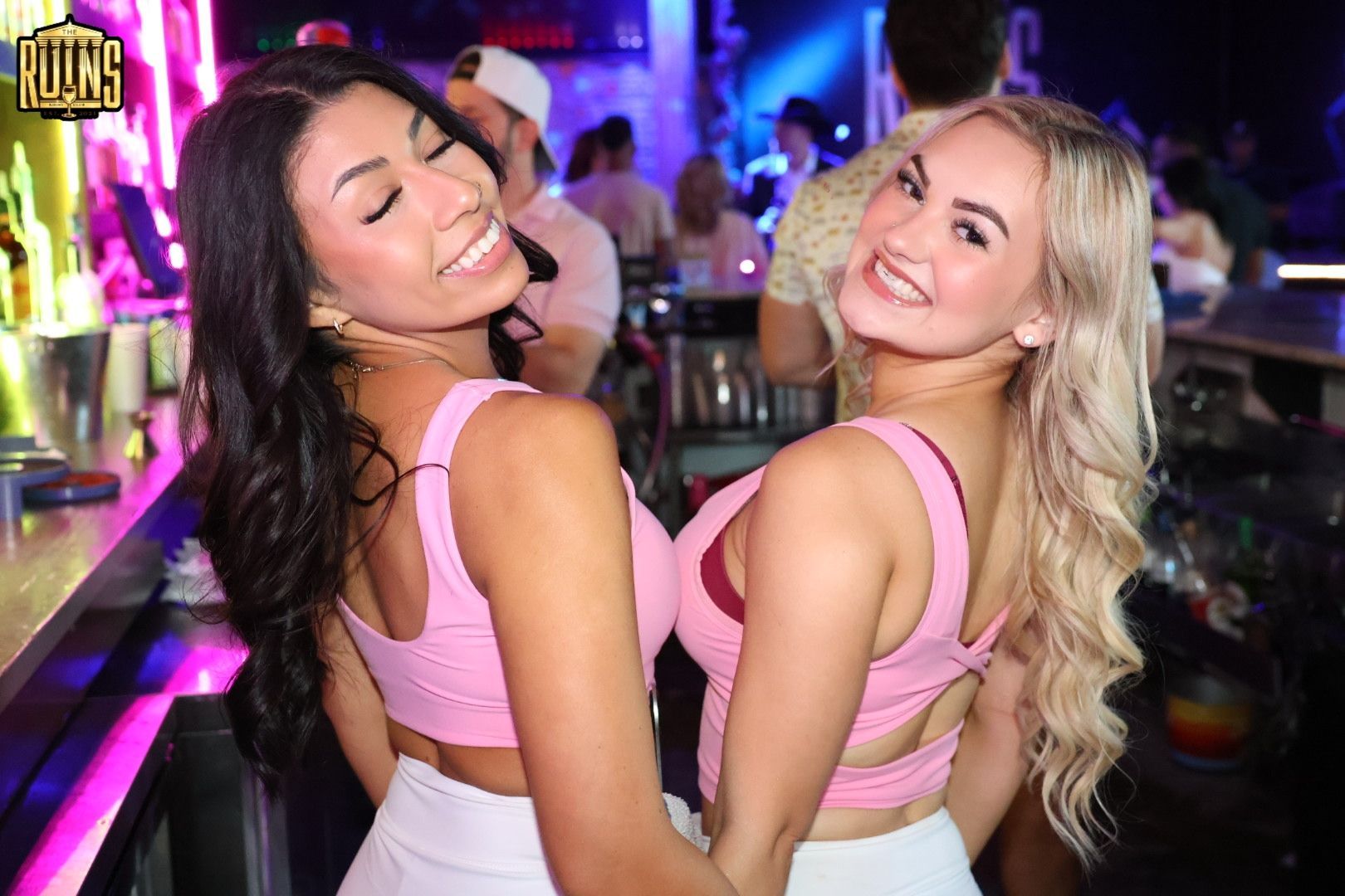 Two women smiling back-to-back at a colorful nightclub bar, wearing pink tops.