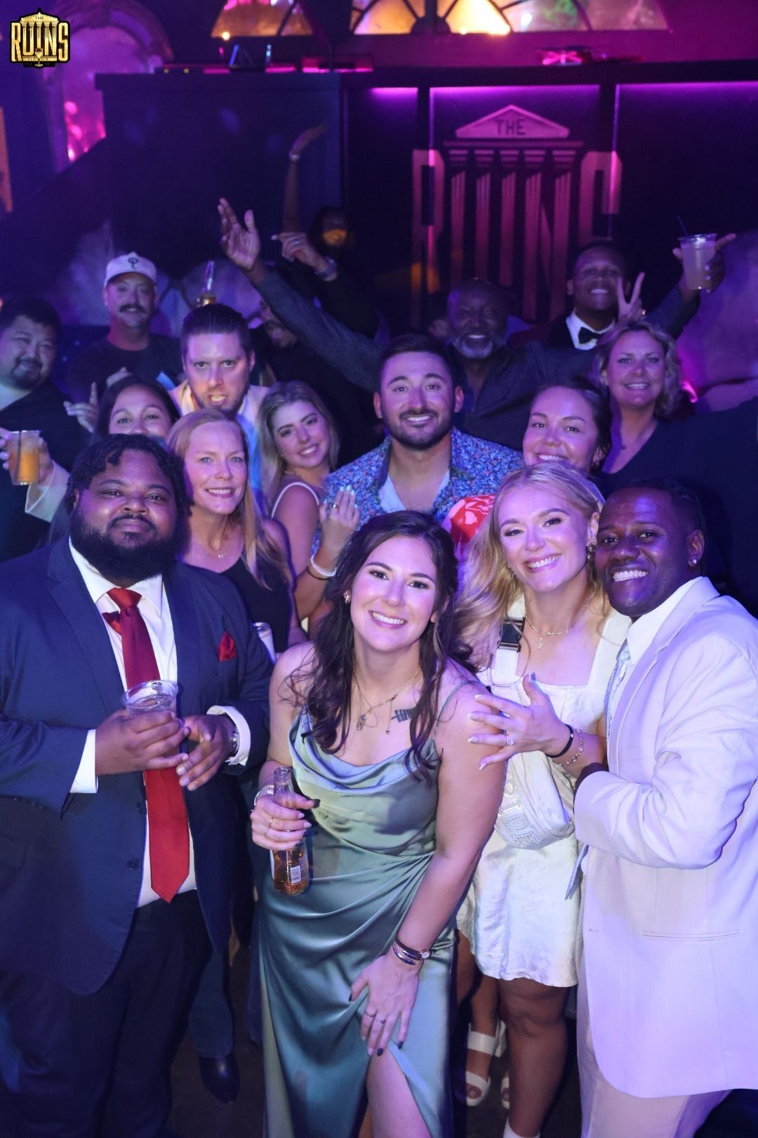 Group of people posing closely together at a dimly lit party with purple lighting and drinks.
