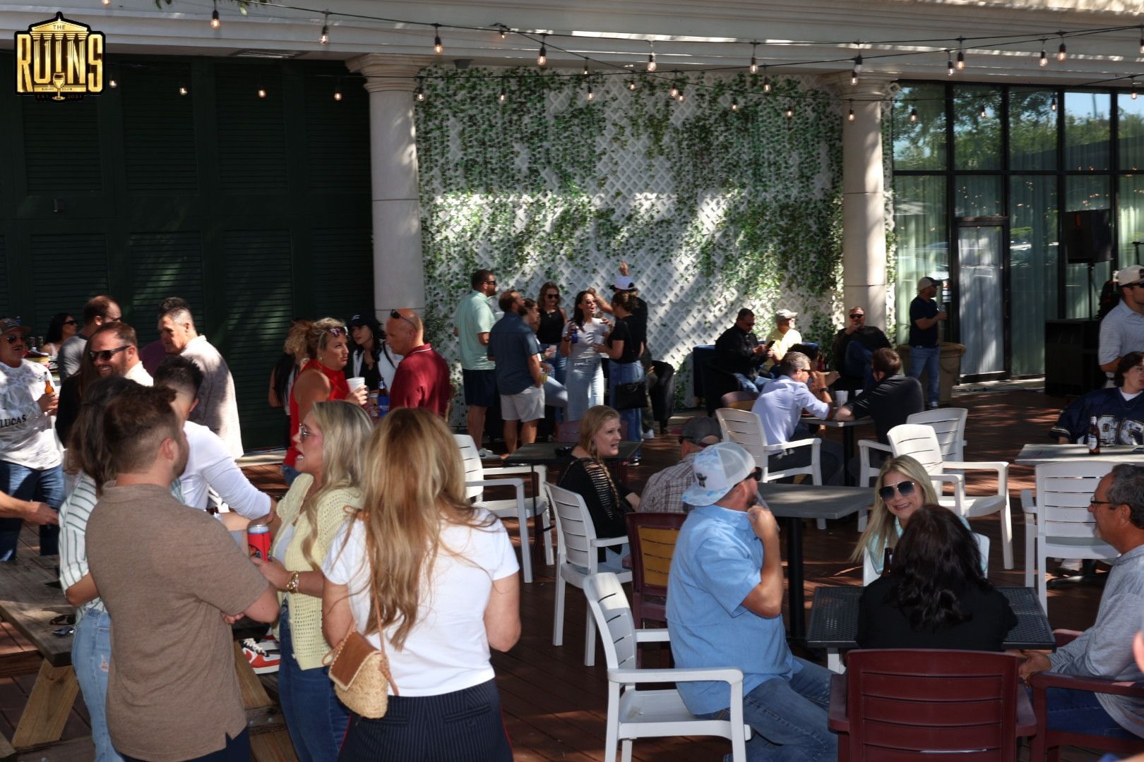 Crowd gathered at outdoor café tables under a shaded patio, with people chatting and standing nearby.