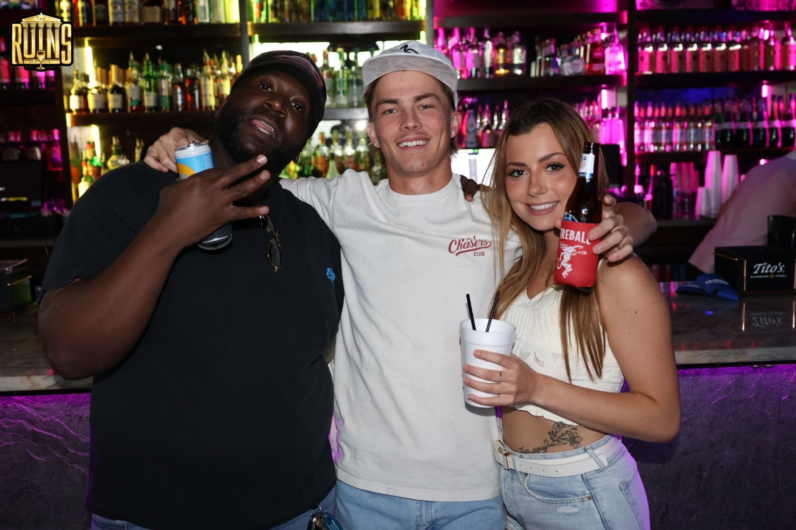Three people smiling at a bar, holding drinks, with colorful bottles glowing in the background.