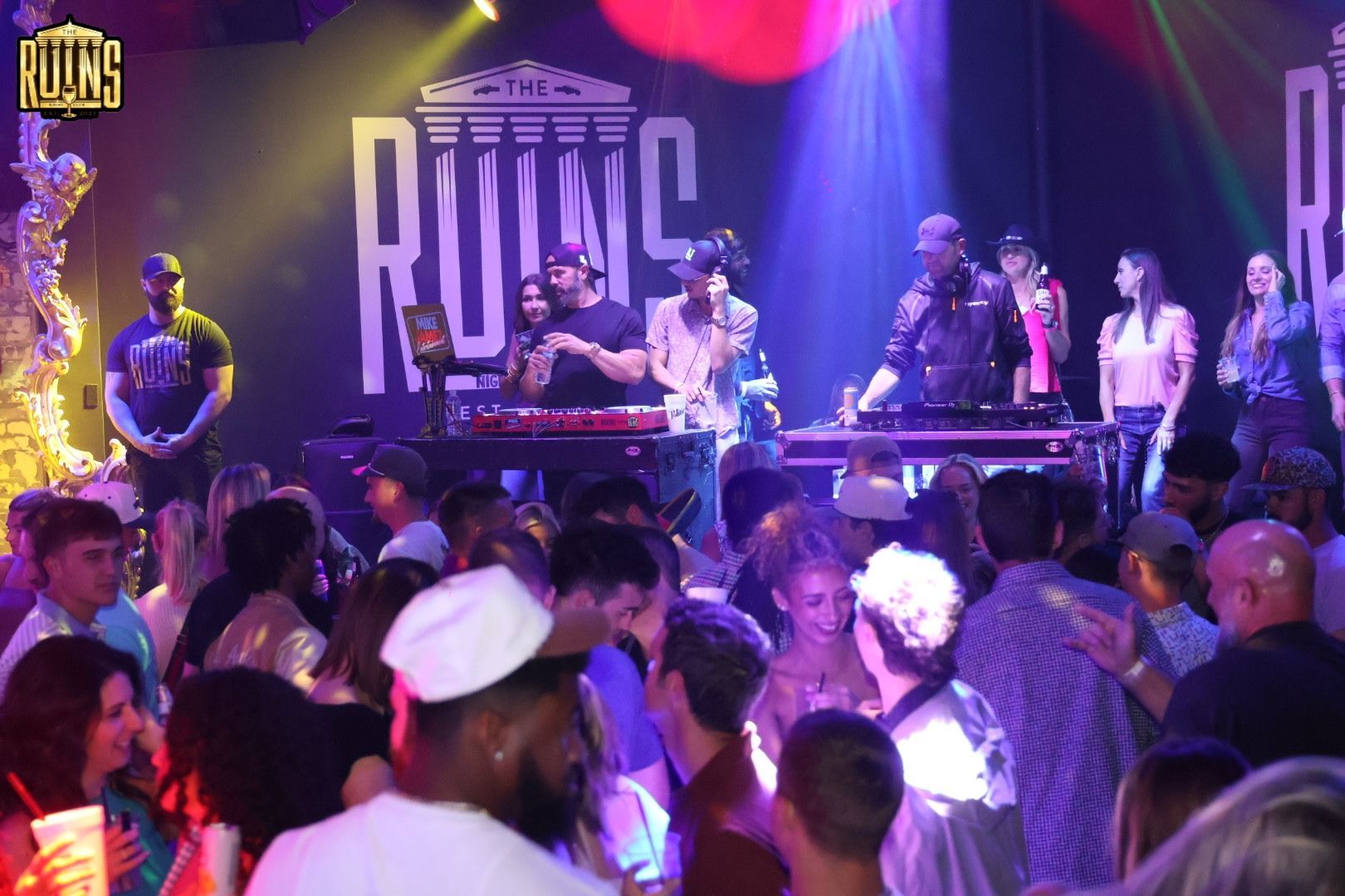 Crowd dancing before a lit stage with performers and purple-pink lights at a club event