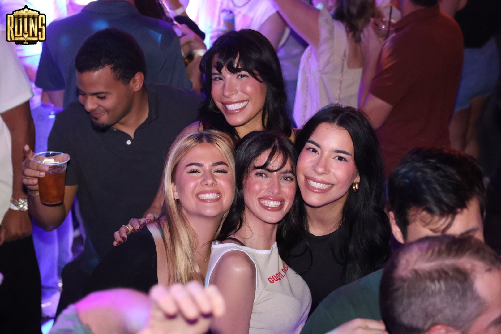 Four friends smiling and posing at a crowded party with purple lighting and drinks nearby