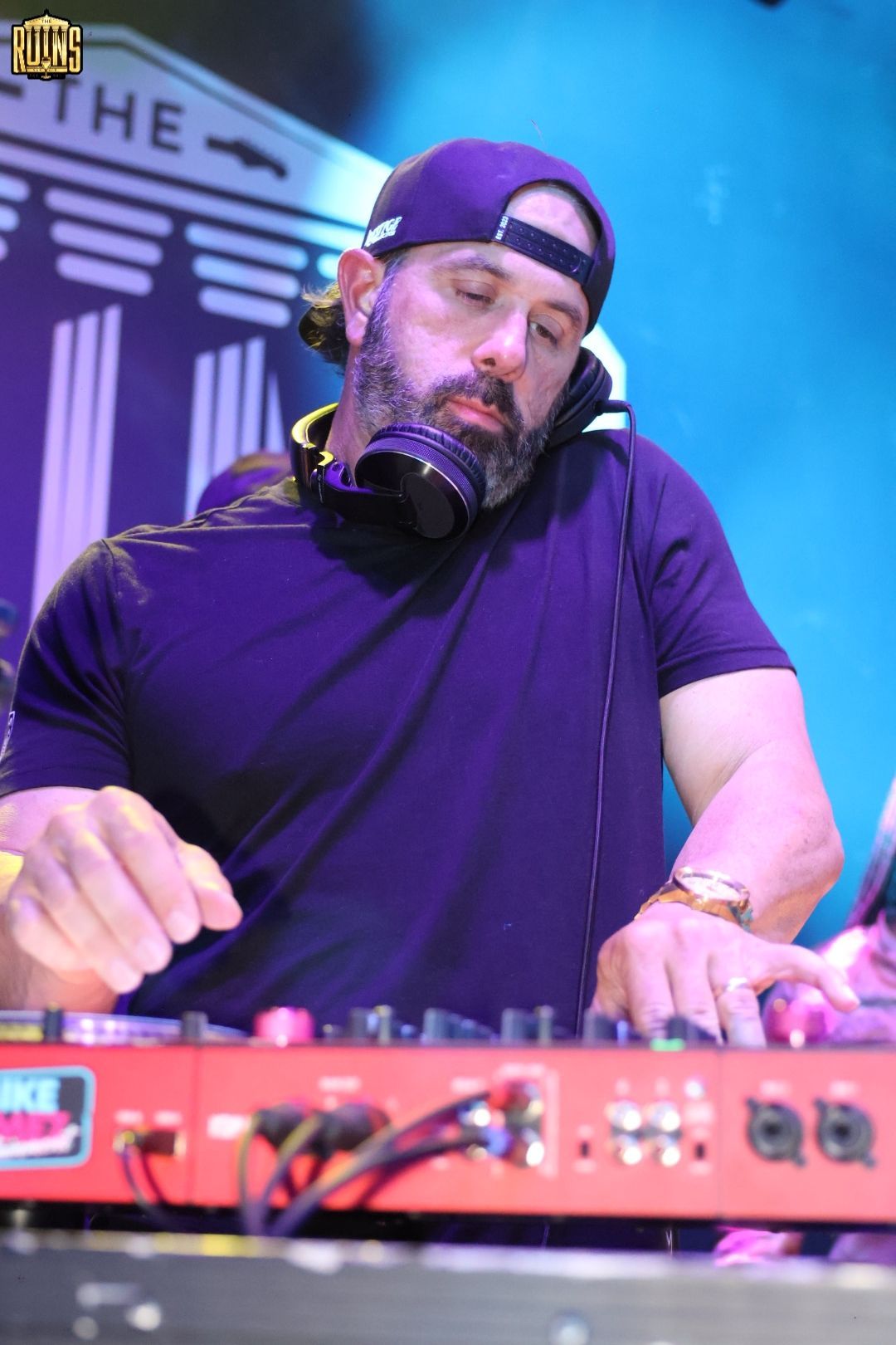 DJ performing at a mixer under blue stage lights, wearing a backward cap and headphones