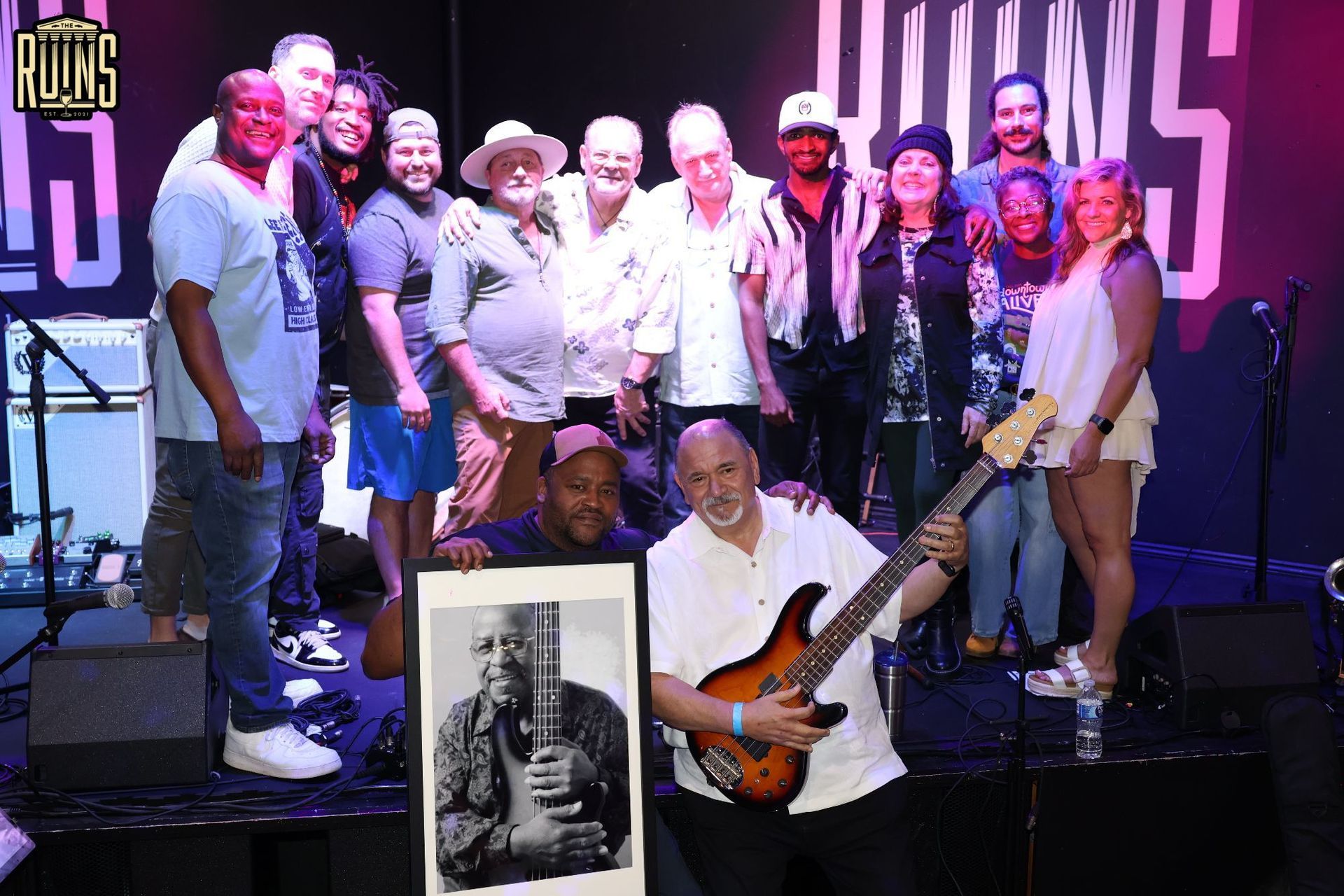 Group of people on stage posing with a guitarist; inset black-and-white portrait at the bottom center.