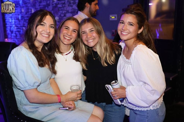 Four people smiling and posing together at a dimly lit party, holding drinks.