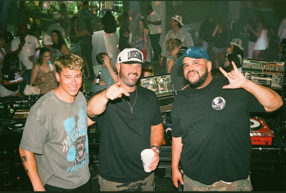 Three smiling people posing at a nighttime car meet, one holding a drink and another flashing a peace sign.