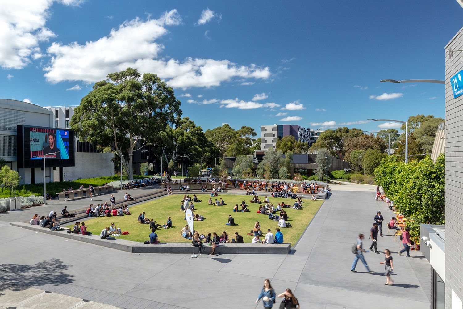 People relax on a grassy square at an outdoor campus. Blue sky, large screen, buildings.