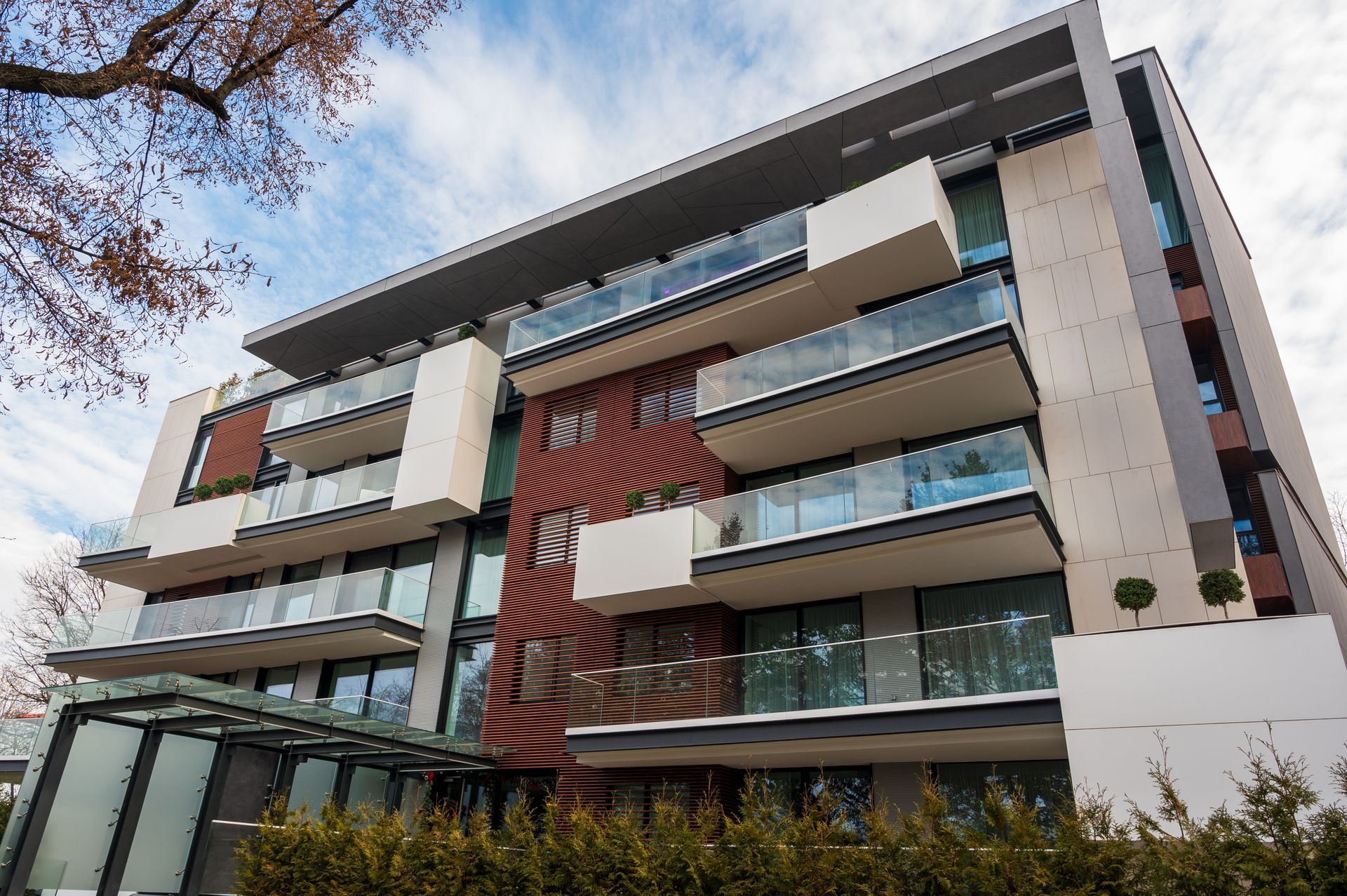 Modern apartment building with glass balconies, white and brown facade, trees in front.