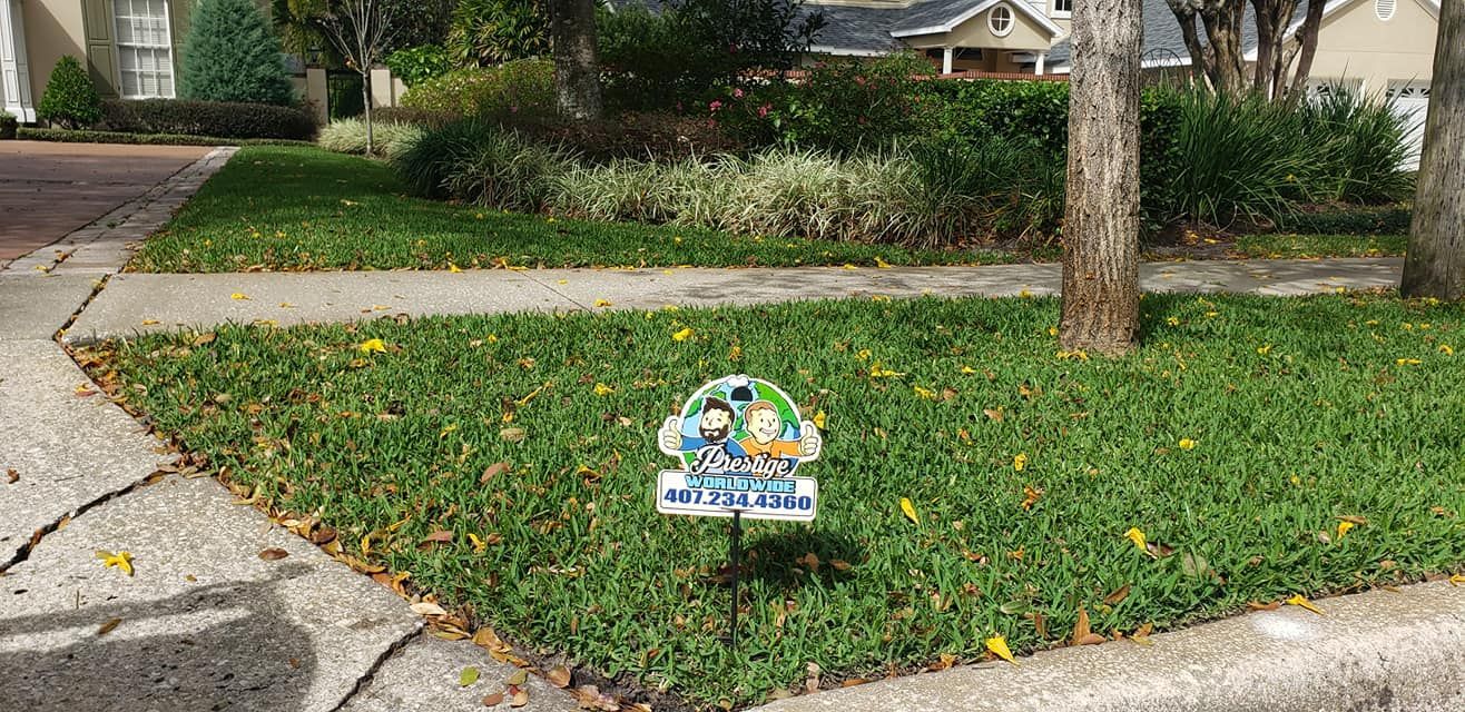 Small yard sign on a grassy lawn beside a sidewalk, in front of a house and tree.