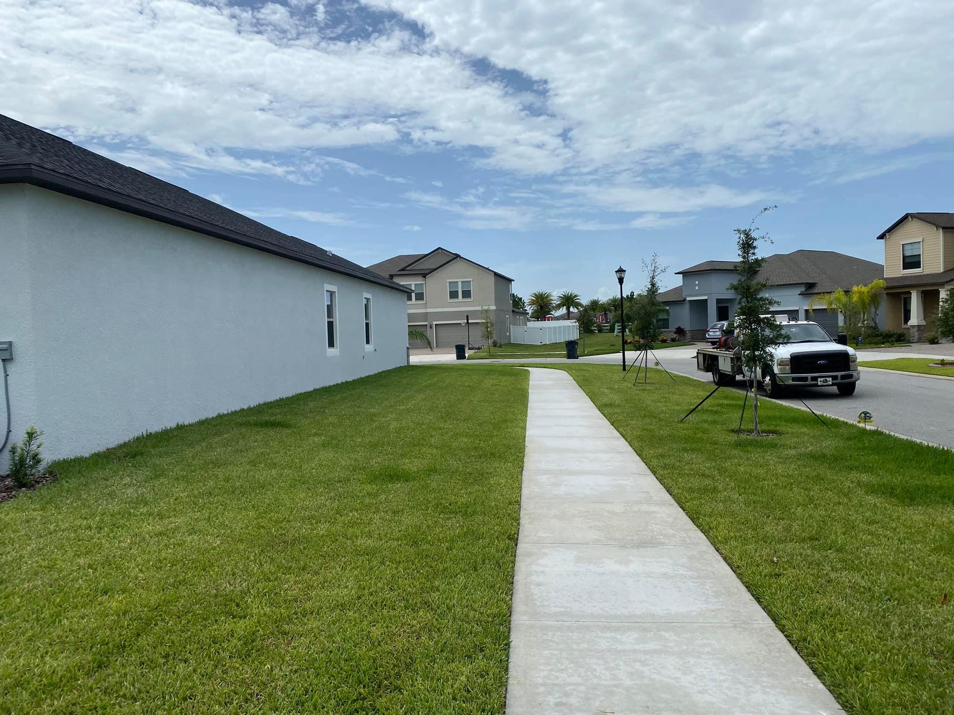 Suburban sidewalk beside white house and green lawn under blue sky, with parked cars and homes in the distance