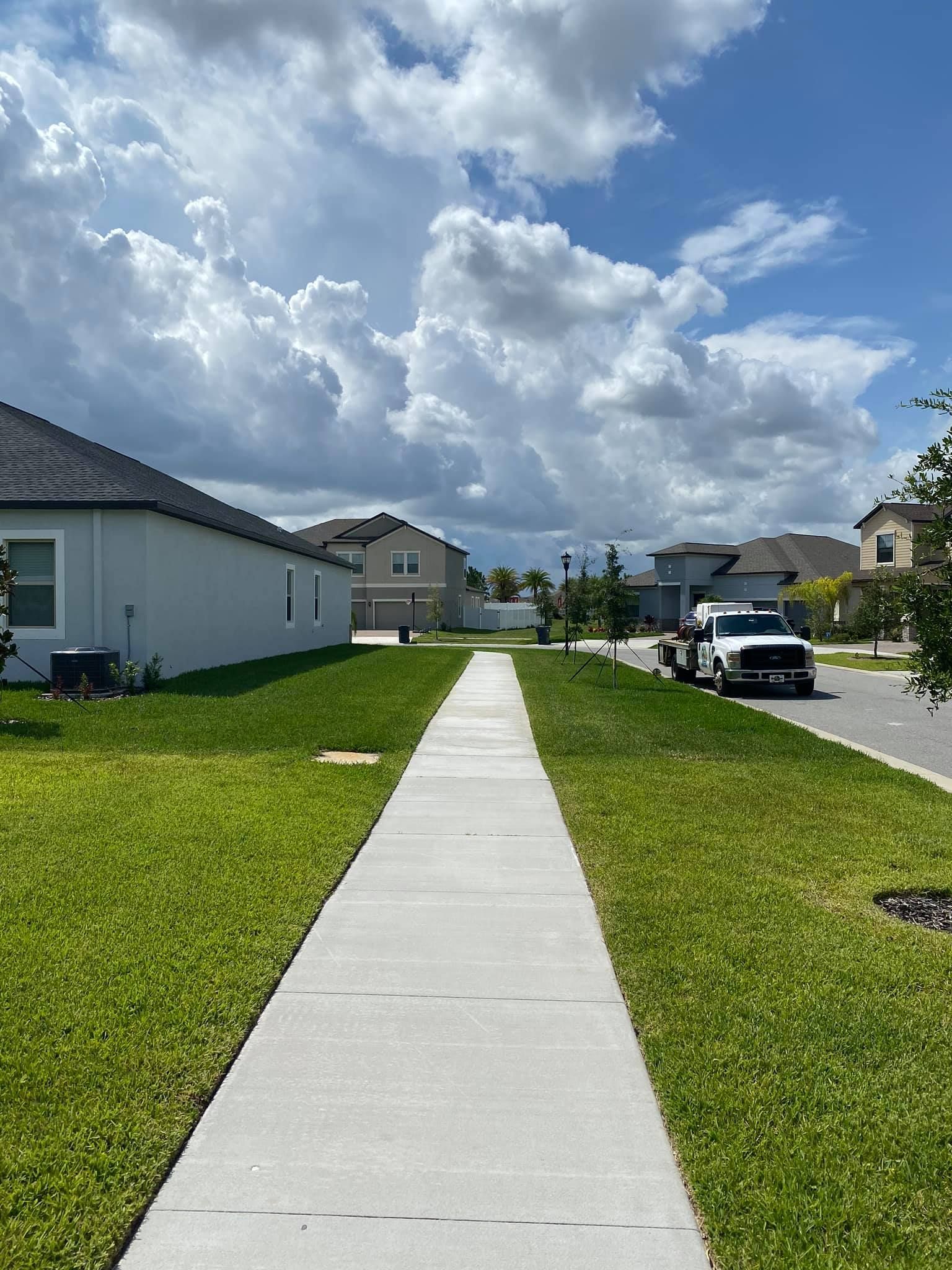 Sidewalk through a suburban neighborhood under a cloudy blue sky, with green lawns and parked cars.