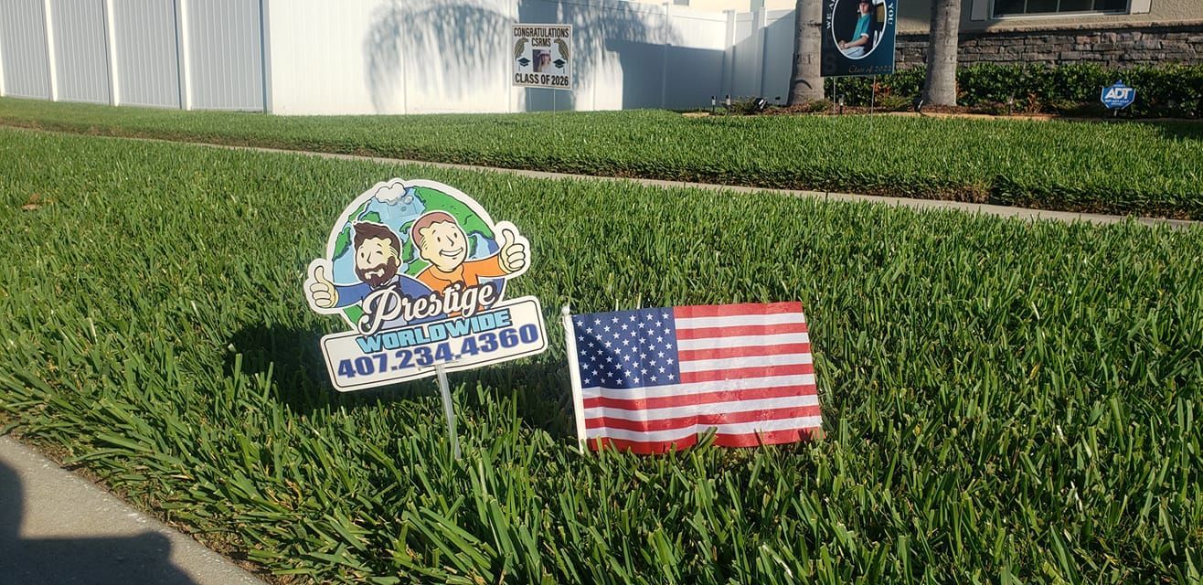 Two small lawn signs in grass, including a waving American flag and a colorful cartoon sign near a sidewalk.