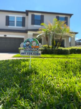 Small garden sign in bright green grass in front of a two-story house with palm trees.
