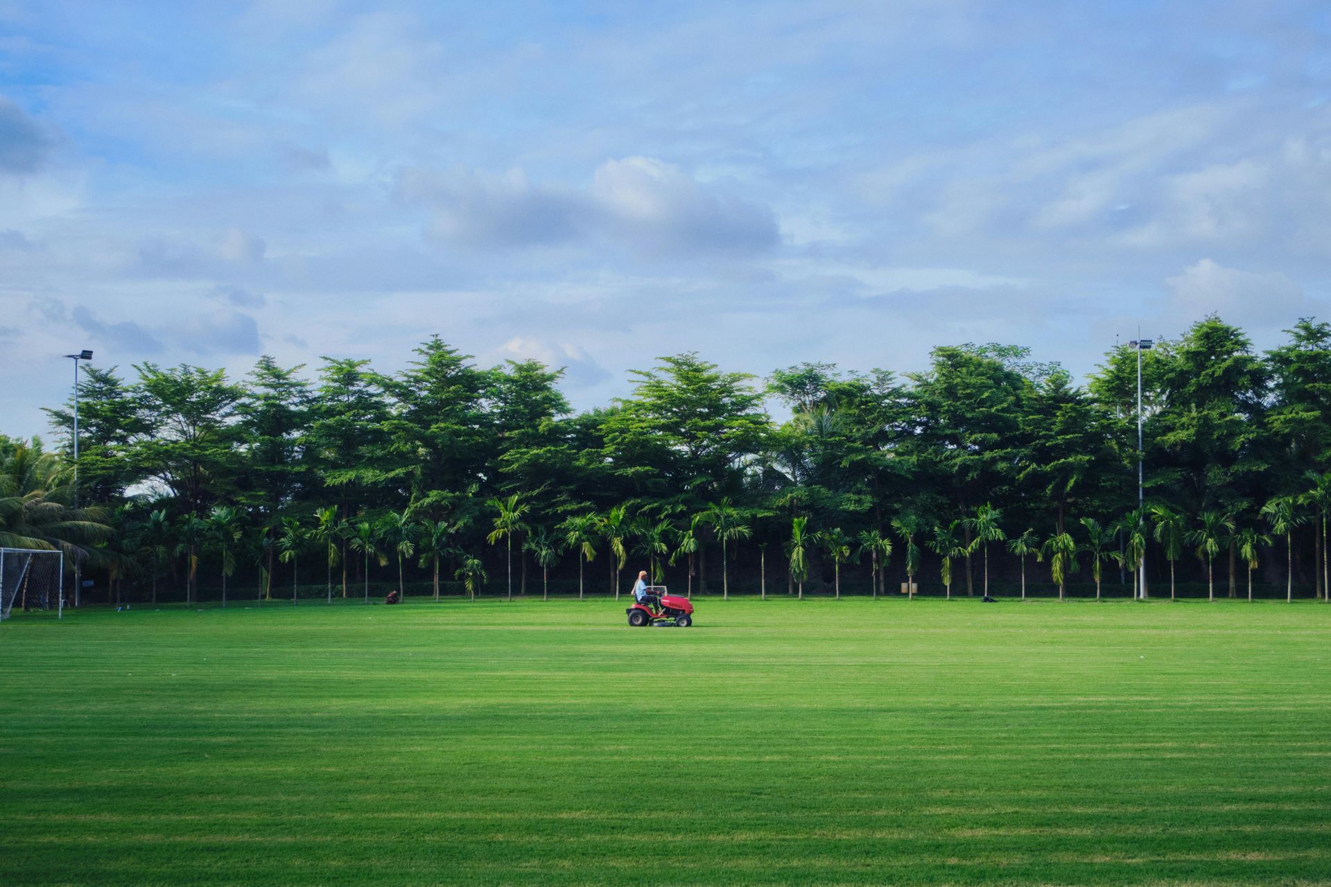 A red tractor on a wide green field with a row of trees under a cloudy blue sky