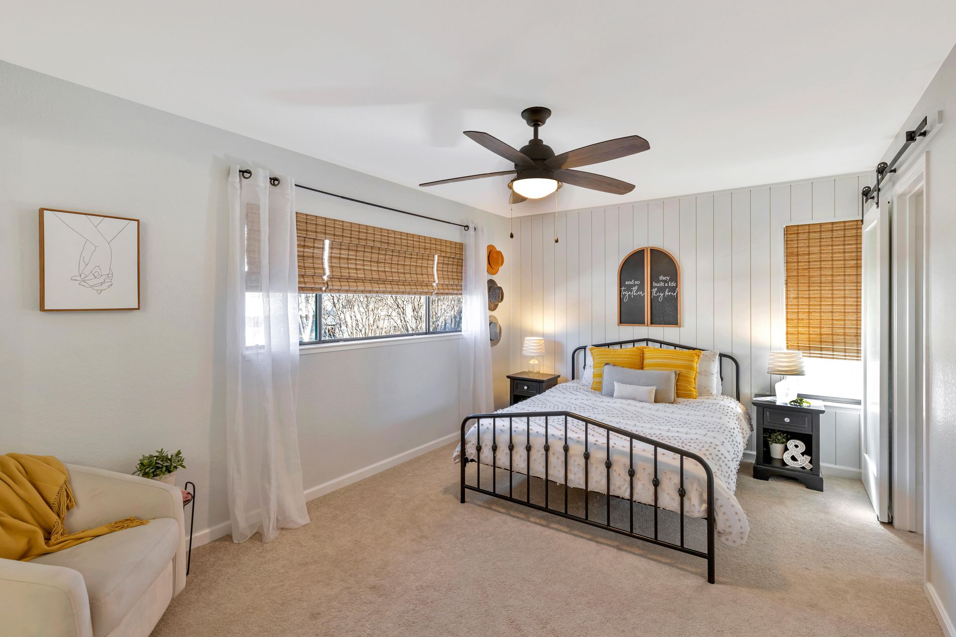 Bedroom with a black bed frame, light-colored walls, ceiling fan, woven blinds, and a small sofa.