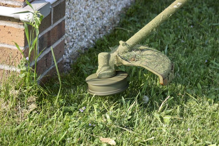 Green garden hose nozzle resting on grass beside a brick wall and white fence post