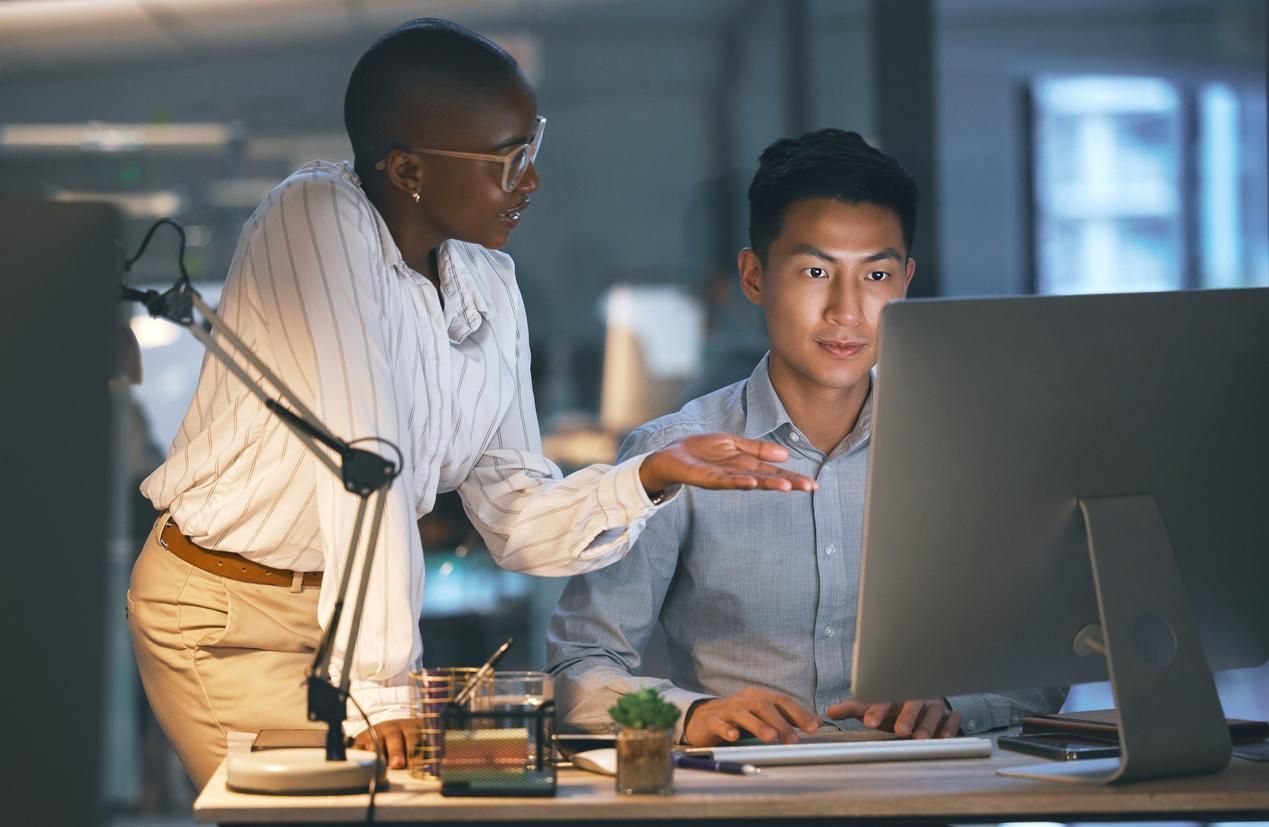 Two coworkers in an office, one pointing at a desktop monitor while the other sits and looks at the screen.