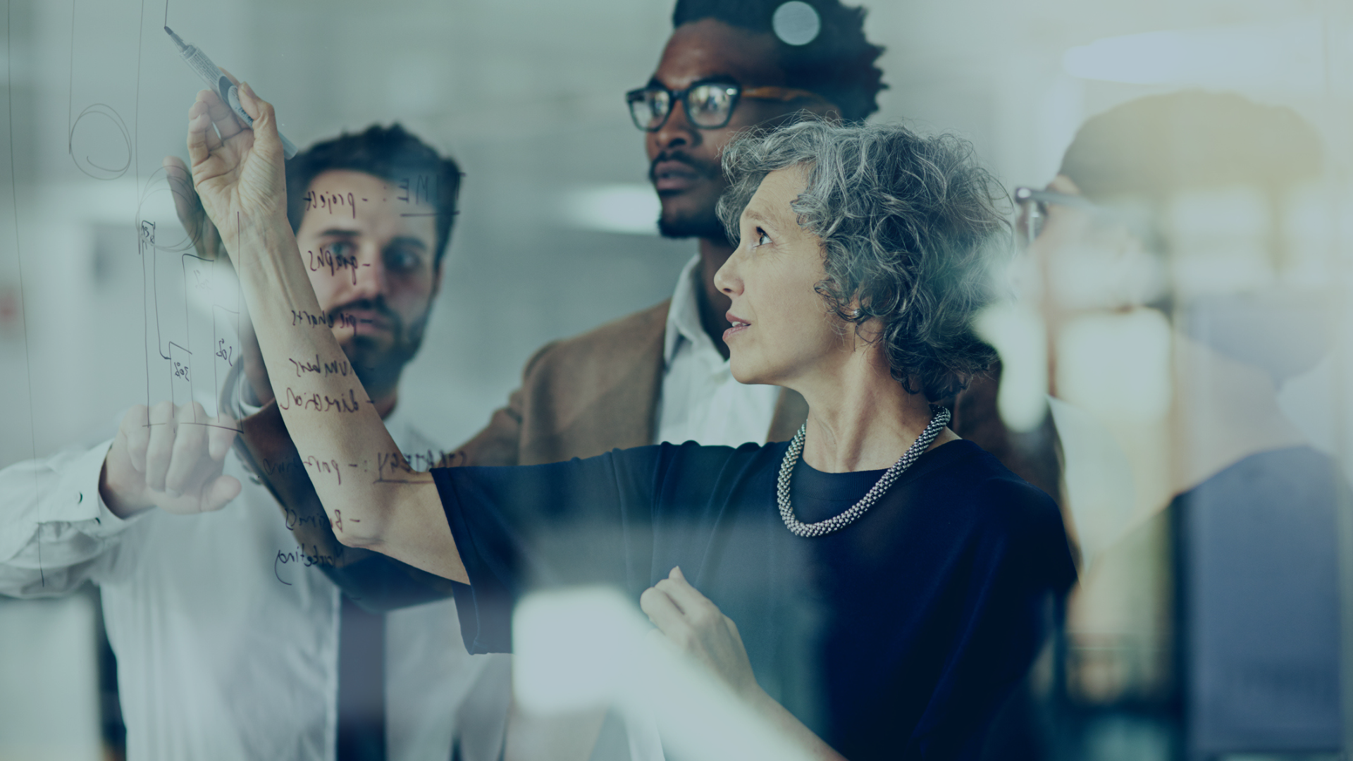 Three coworkers discuss notes on a glass wall in a bright office.