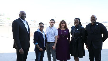 Six people standing outdoors, posing together and smiling in formal attire.