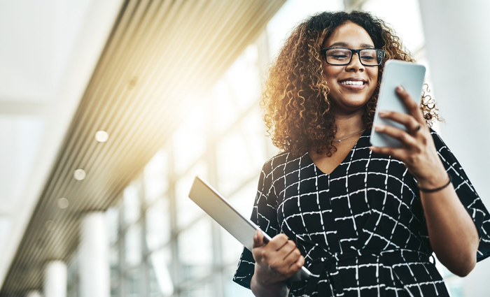 Smiling person holding a phone and tablet in a bright office hallway