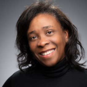 Smiling person with dark wavy hair and black turtleneck against a gray studio background