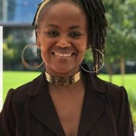 Smiling person in a black shirt and gold choker necklace standing outdoors with blurred buildings behind them