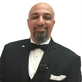 Man in a black tuxedo with bow tie and pocket square, smiling against a white background