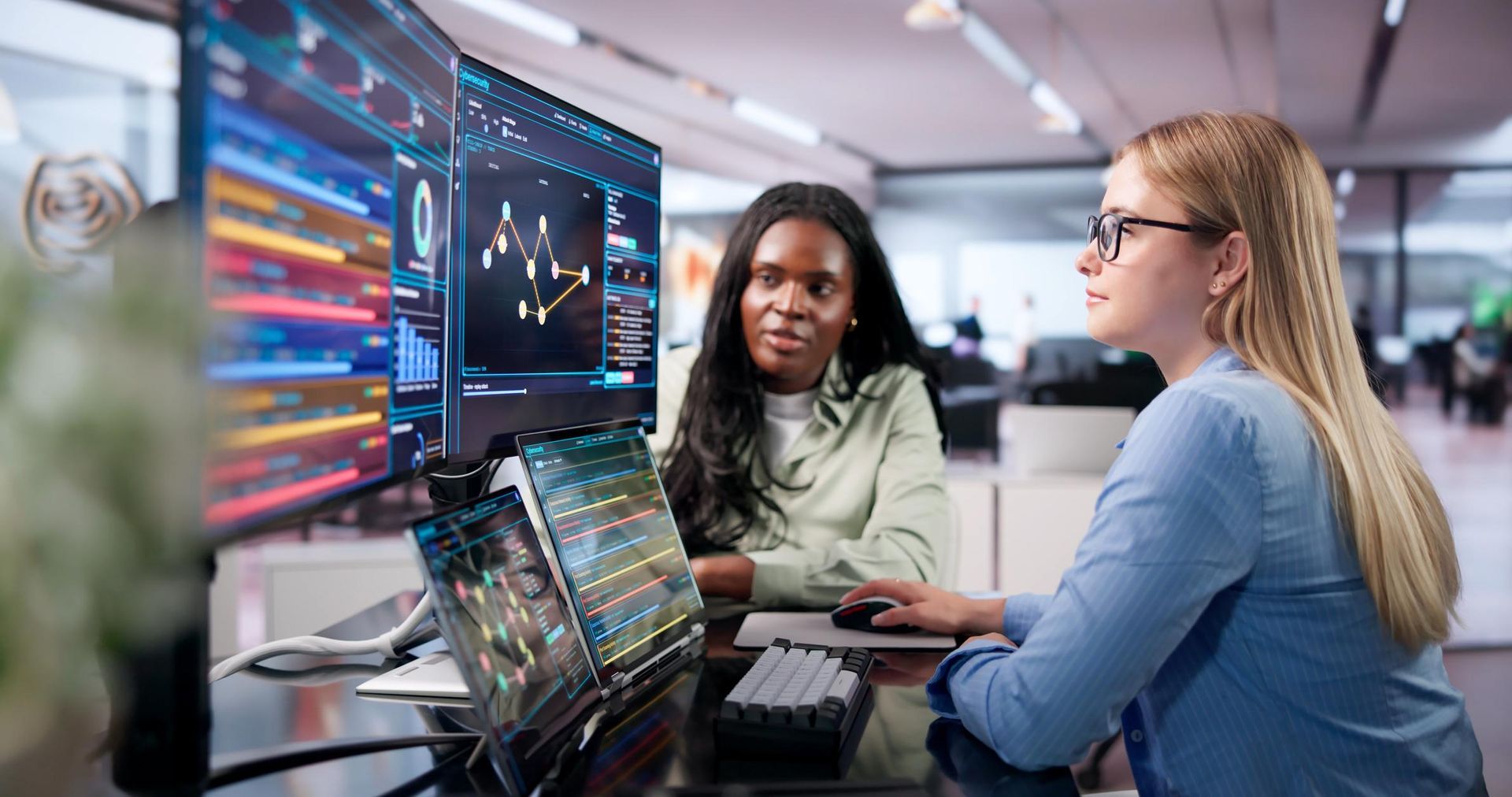 Two coworkers reviewing data on laptops in a modern office, with colorful charts on a large monitor.