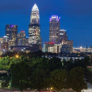 City skyline at dusk with illuminated high-rises above a dark green treeline.
