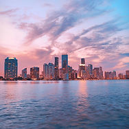 City skyline at sunset reflected on calm water, with pink clouds over tall buildings.