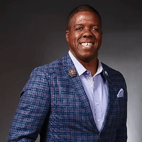 Smiling man in a blue plaid blazer and light shirt against a dark studio background