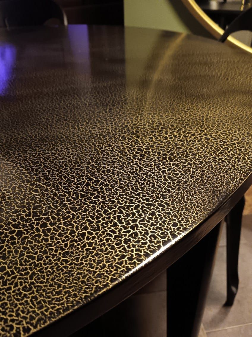 Tavolo rotondo scuro con superficie texturizzata e punteggiata d'oro, in un ambiente interno con luci soffuse.