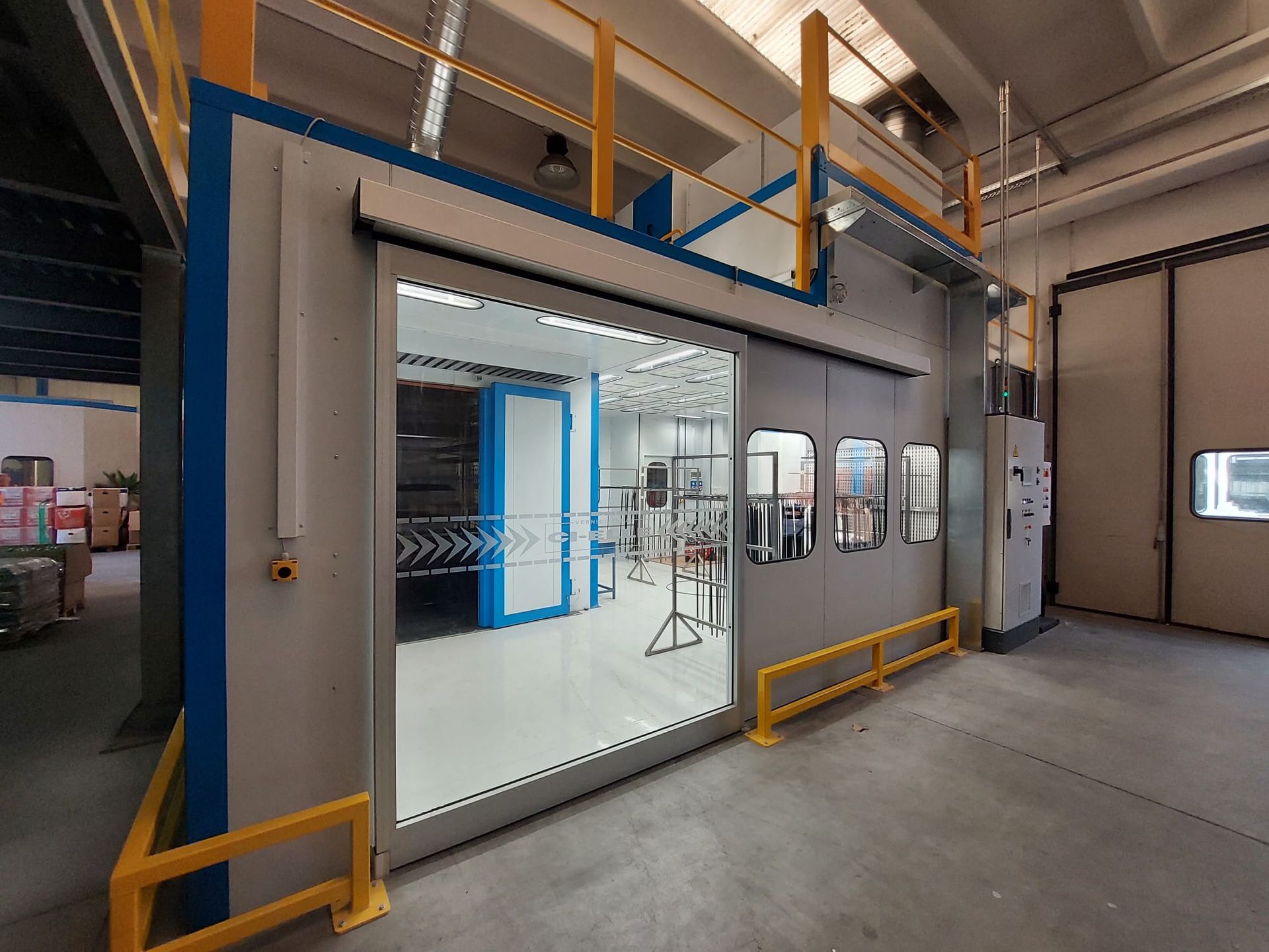Cabina di verniciatura industriale con grande porta scorrevole bianca, pareti grigie e ringhiere di sicurezza gialle all'interno di una fabbrica.