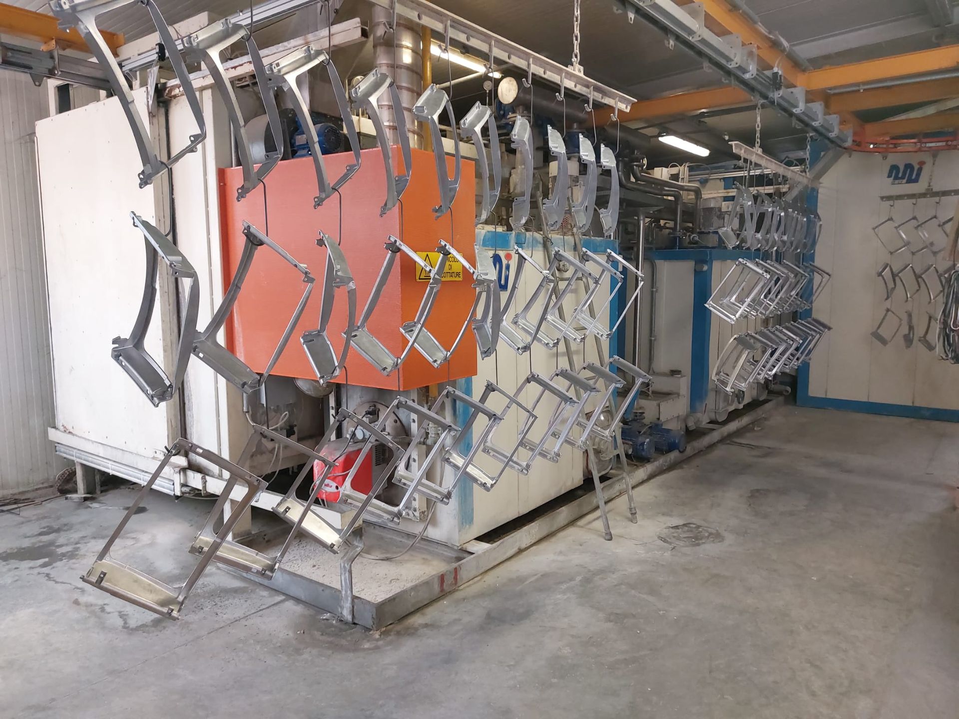 Macchinari industriali con telai metallici sospesi e pannello centrale arancione in un ambiente di fabbrica.