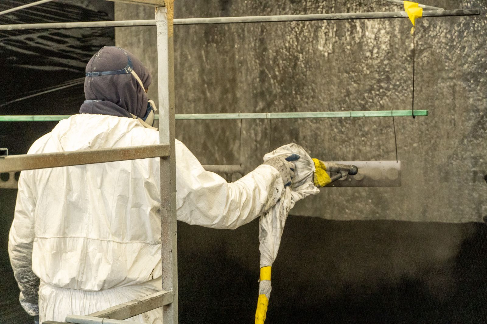 Un operaio in tuta bianca vernicia a spruzzo un muro di cemento da una piattaforma di ponteggio.